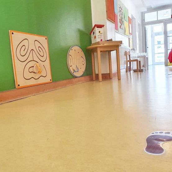 Ein Flur mit grünen Wänden, einem Holztisch mit einem Vogelhäuschen, einem Fußaufkleber und einem kleinen Stuhl. Es gibt farbenfrohe Gemälde an der Wand und eine Glastür zu einem anderen Raum.