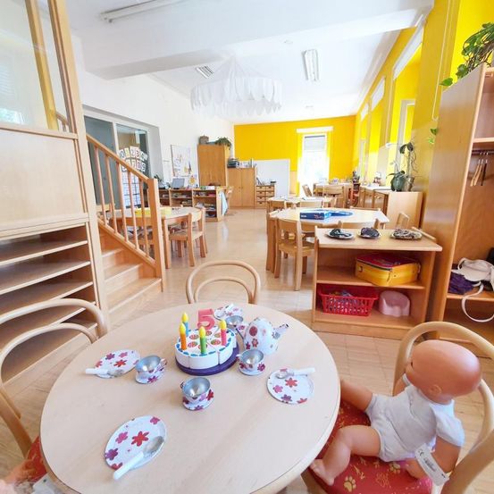 Ein Spielzimmer mit gelben Wänden und Holzmöbeln. Ein Tisch hat einen Geburtstagskuchen, Teller, Tassen und eine Teekanne. Eine Babypuppe sitzt auf einem Stuhl. Es gibt Regale und eine Treppe im Raum.