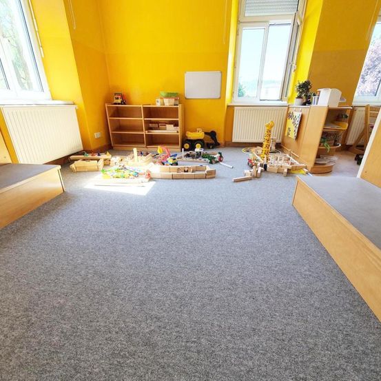 Ein Spielzimmer mit gelben Wänden, Teppichboden und verstreuten Spielzeugen. Holzregale, ein Heizkörper und eine Tafel sind vorhanden. Fenster mit Jalousien lassen natürliches Licht herein.