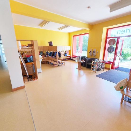 Ein heller, leerer Raum mit gelben Wänden und weißem Boden. Es gibt Regale und Stühle mit Taschen. Eine Tür mit dem Wort 'mein' ist offen.