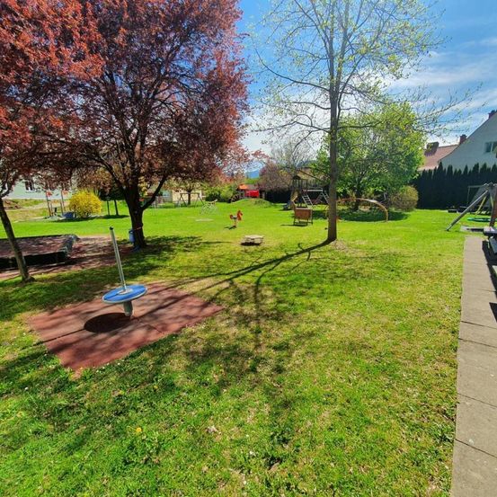 Ein Spielplatz mit Schaukel und Wippe auf einem sattgrünen Rasen unter einem blauen Himmel mit verstreuten Wolken. Bäume, ein Haus und ein Zaun sind in der Ferne.