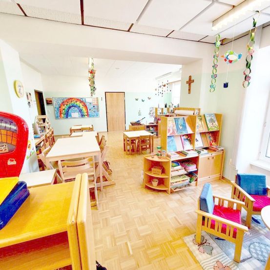 Ein Klassenzimmer mit Holztischen und Stühlen. Ein Bücherregal voller Bücher und ein Regenbogenbild schmücken die Wand. Bunte Dekorationen hängen von der Decke.