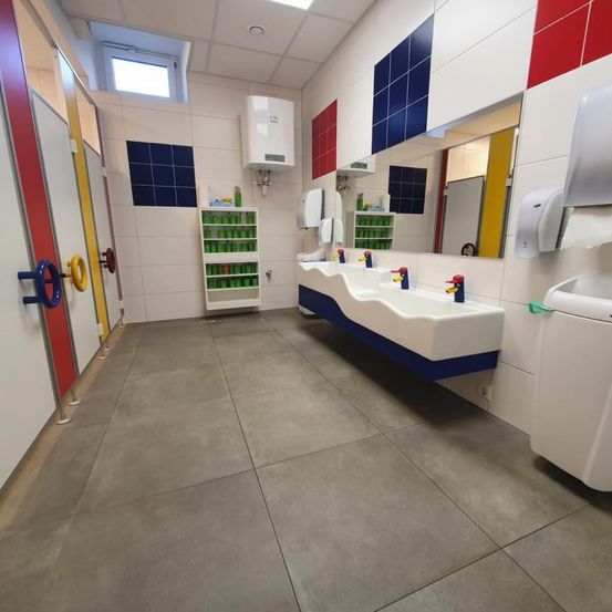 Ein sauberes Badezimmer mit gefliesten Wänden, drei Waschbecken mit Seifenspendern, einem Spiegel und einem Wasserkocher. Bunte Fliesen schmücken die Wände.