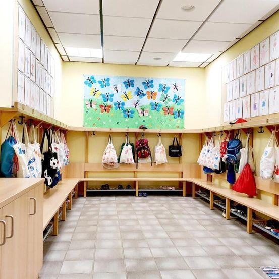 Ein Schulumkleideraum mit Regalen, die Taschen und Schuhe halten. Ein Schmetterlingsbild hängt an der Wand und Bilder von Kindern schmücken die Wände. Der Raum hat Holzbänke und gefliesten Boden.