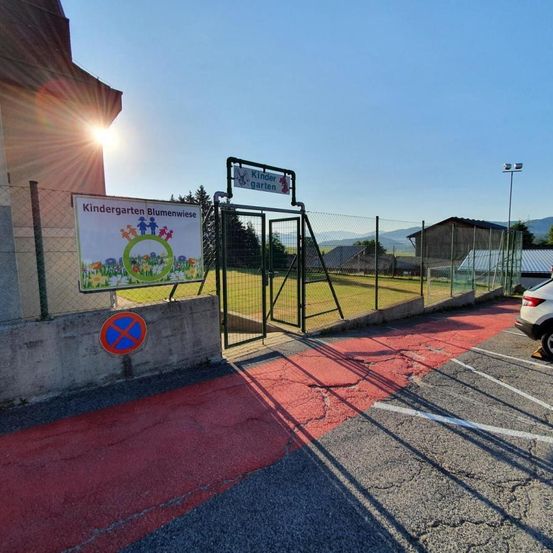 Ein Kindergarteneingang mit einem Schild, das 'Kindergarten Blumenwiese' lautet. Ein roter Weg führt zu einem geschlossenen Tor, und ein Gebäude ist im Hintergrund zu sehen.