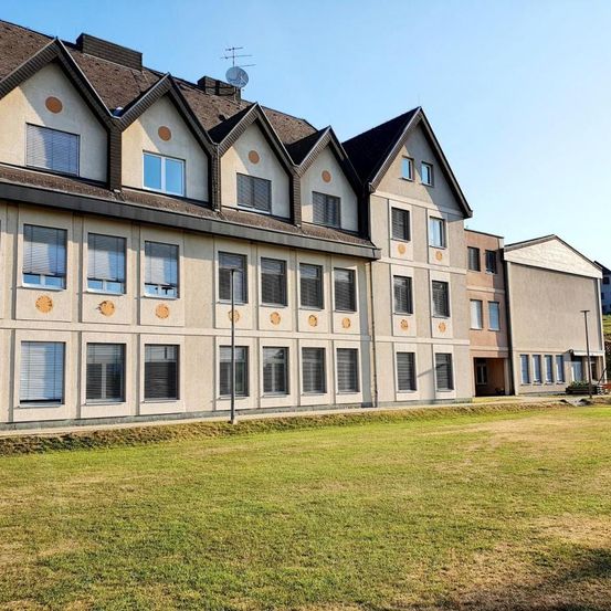 Ein mehrstöckiges Gebäude mit dreieckigem Dach steht vor einer Wiese unter klarem Himmel.