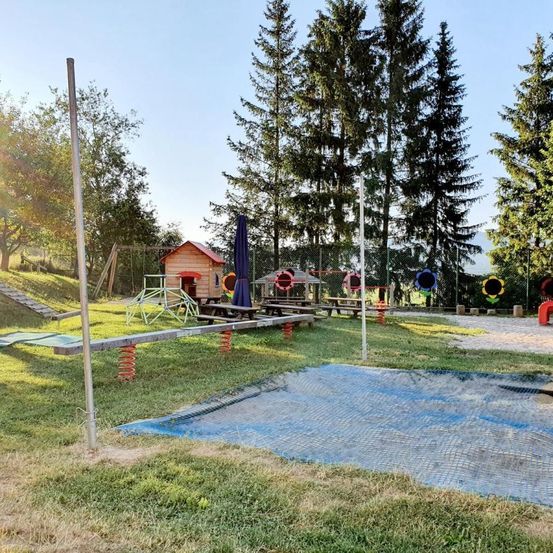 Ein Freispielplatz mit verschiedenen Spielgeräten, einer Schaukel und einem kleinen Holzhaus. Es gibt auch Picknicktische und einen geschlossenen Schirm. Der Bereich ist von Bäumen umgeben.