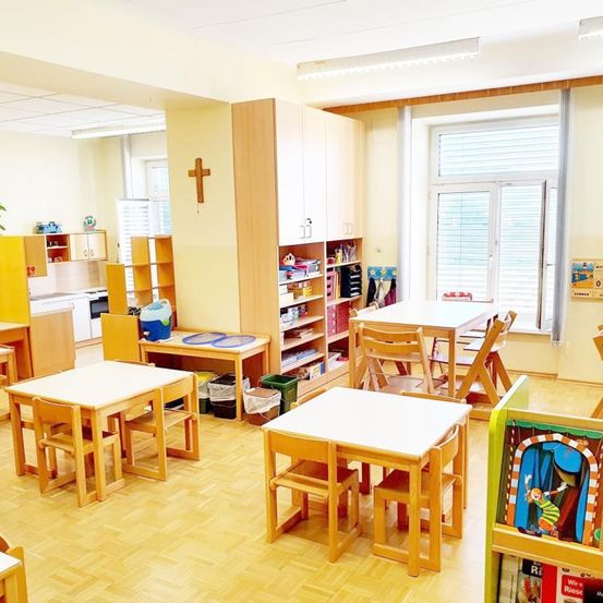 Ein Klassenzimmer mit Holzmöbeln, Tischen, Stühlen und einem Schrank. Ein Kreuz an der Wand und ein Fenster mit Jalousien in der Nähe.