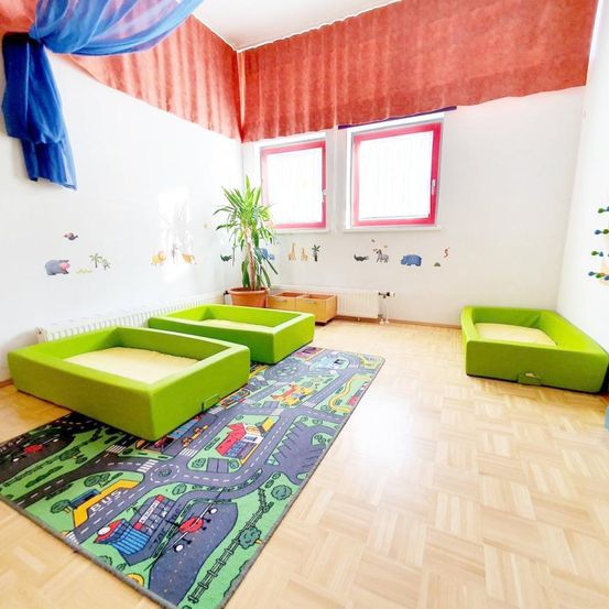 Ein Spielzimmer mit Holzboden hat grüne Sitzsäcke und einen Spielteppich. Es gibt Fenster, einen Heizkörper und eine Wand mit Tieraufklebern.