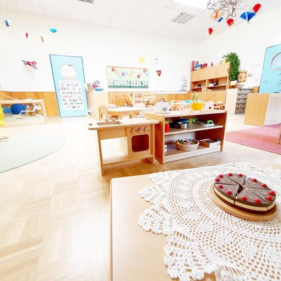 Ein hell beleuchteter Raum mit Holzboden, ausgestattet mit einem Tisch, auf dem ein Kuchen auf einem Doily steht, Holzmöbeln und bunten Dekorationen.