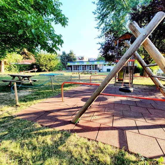 Ein Spielplatz mit Schaukeln, einer Rutsche und Picknicktischen unter Bäumen. Im Hintergrund steht ein Gebäude mit einem roten Dach.