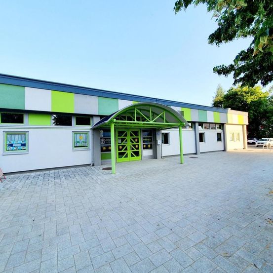 Ein Gebäude mit einer grün-weiß gestreiften Fassade und einem grünen Eingangsvordach. Im Hintergrund sind eine Bank und einige Bäume zu sehen.