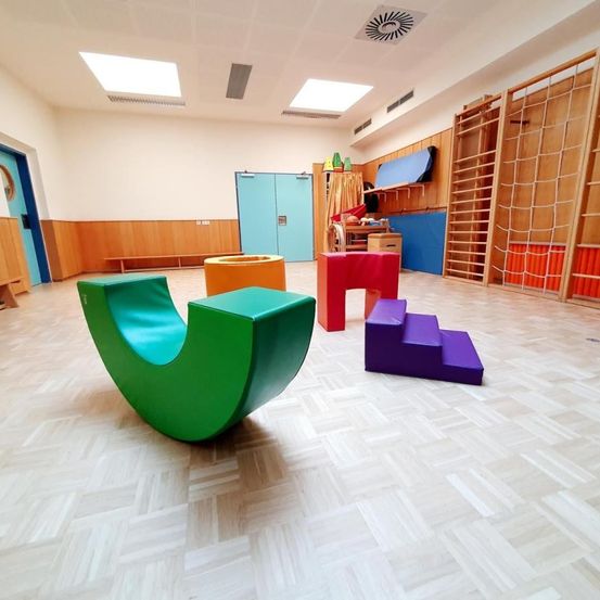 Ein heller Raum mit Holzwänden und einem weißen Holzboden. Es gibt bunte Stühle, eine Bank und Kletterstrukturen. Eine Tür mit einem runden Spiegel befindet sich auf der linken Seite.