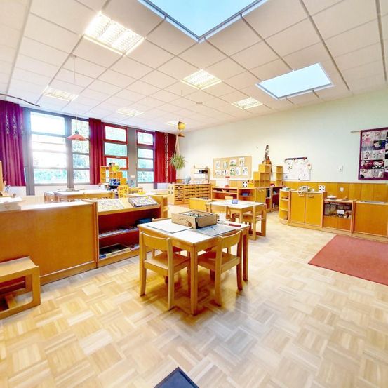 Ein Klassenzimmer mit Holztischen und -stühlen, Regalen voller Bücher, einem roten Teppich und großen Fenstern mit roten Vorhängen.