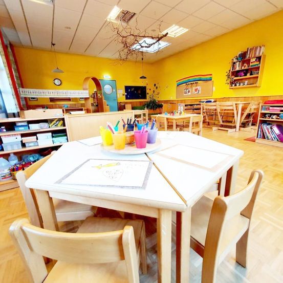 Ein hell beleuchteter Klassenraum mit gelben Wänden, Holzstühlen und Tischen. Kunstzubehör liegt auf dem Tisch, und Regale mit Büchern und Gegenständen befinden sich an der Seite.