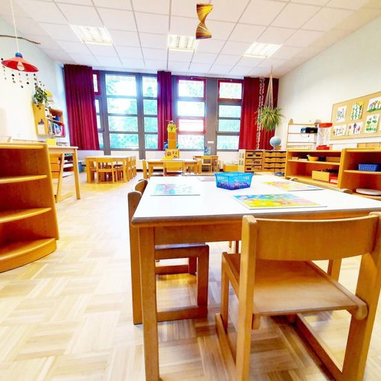 Ein Klassenzimmer mit Holztischen und Stühlen um einen langen Tisch mit Büchern und Spielzeug herum angeordnet. Der Raum hat weiße Wände, Holzböden und große Fenster mit roten Vorhängen.