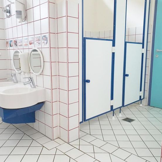 Ein sauberes Badezimmer mit weißen gefliesten Wänden und blauen Akzenten. Es hat ein Waschbecken, zwei Spiegel und drei Duschkabinen. Der Boden ist weiß und sauber.