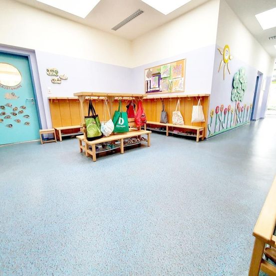 Ein leerer Flur mit blauem Teppichboden. Holzbänke mit befestigten Schuhregalen. Taschen hängen an Haken. Eine runde blaue Tür mit einem Spiegel. Eine Wand mit bunten Zeichnungen.