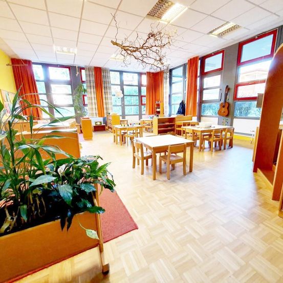 Ein Klassenzimmer mit Holztischen, Stühlen und großen Fenstern. Eingepflanzte Pflanzen und eine Gitarre schmücken den Raum. Der Raum hat Holzboden und Deckenleuchten.