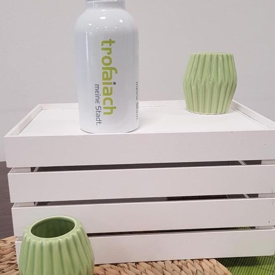 Eine weiße Flasche mit 'trofalach meine stadt' Text und einem schwarzen Deckel auf einer weißen Holzkiste. Zwei grüne Keramikvasen sind oben und vor der Kiste platziert.