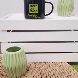 Eine schwarze Tasse mit der Aufschrift 'Stolz der Stadt!' steht auf einem weißen Regal neben zwei grünen Vasen. Die Tasse und die Vasen liegen auf einem gewebten Teppich.
