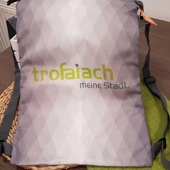 Eine grau-weiße Tragetasche mit geometrischem Muster, beschriftet mit 'trofaiach meine Stadt' in Grün. Sie hat einen schwarzen Gurt und steht auf einem braunen Tisch mit einer gewebten Tischdecke.
