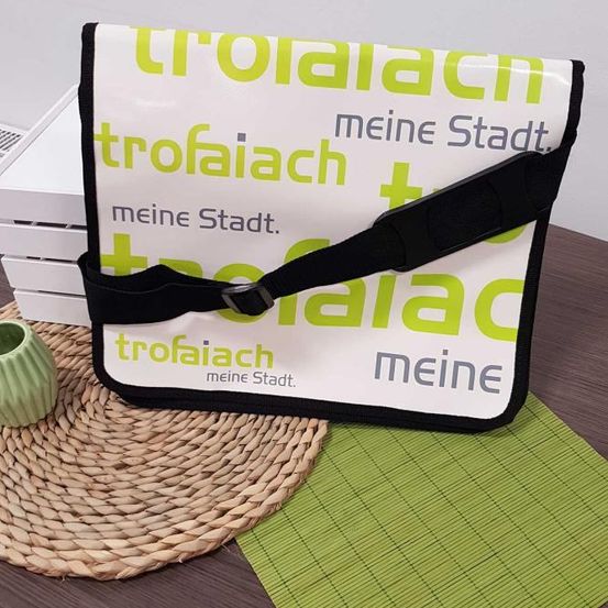 Eine Tasche mit einem grünen und weißen Design, auf der 'trofaiaach meine Stadt' steht, liegt auf einem braunen Tisch. Sie hat einen schwarzen Gurt und befindet sich auf einer geflochtenen Matte.