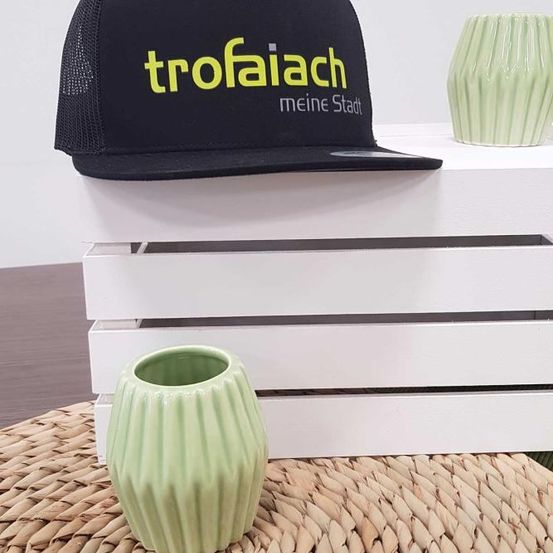 Ein schwarzer Hut mit 'trofaiach' und 'meine Stadt' in Gelb auf einem weißen Holzkasten mit einer grünen Vase auf einem geflochtenen Teppich.