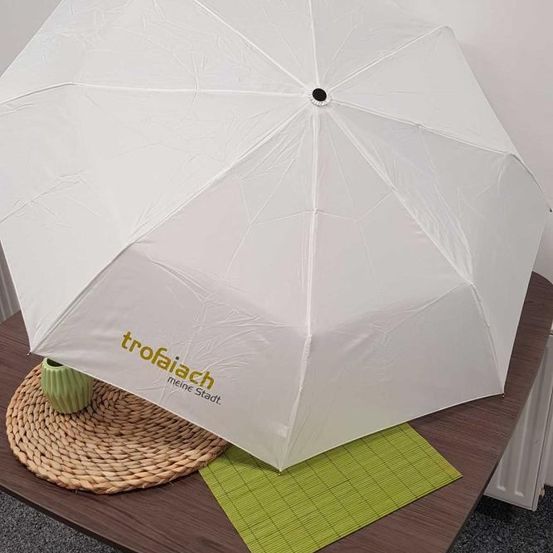 Ein weißer gefalteter Regenschirm mit grünem Text auf dem Baldachin, auf einem braunen Tisch platziert. Der Tisch ist mit einer geflochtenen Unterlage und einem grünen Platzset abgedeckt. Eine grüne Vase steht auf dem Platzset.