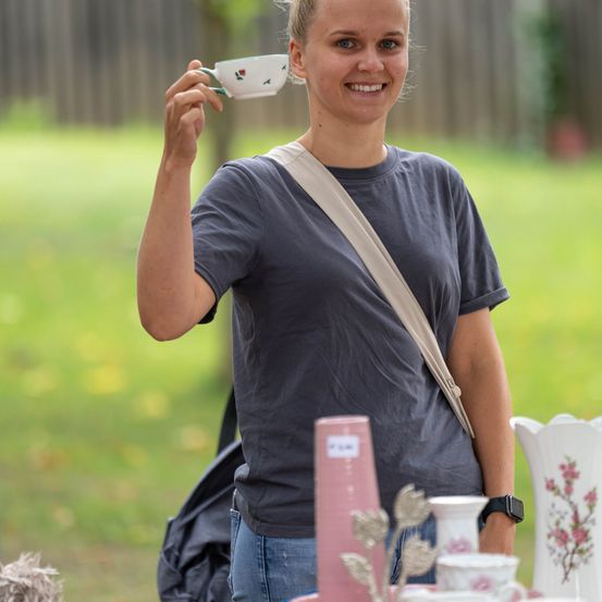 Eine Frau in einem grauen T-Shirt hält eine kleine, blumige Tasse in der Hand und lächelt, mit einem Tisch voller Keramikartikel im Hintergrund.