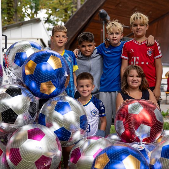 Eine Gruppe von Kindern, Jungen und Mädchen, steht hinter einem Haufen Fußballbälle, einige in Plastik verpackt. Sie lächeln und posieren für ein Foto.
