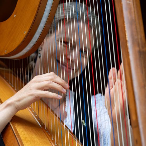 Eine Frau mit grauem Haar spielt Harfe, lächelt und schaut in die Kamera. Sie trägt ein weißes Oberteil und eine schwarze Weste.