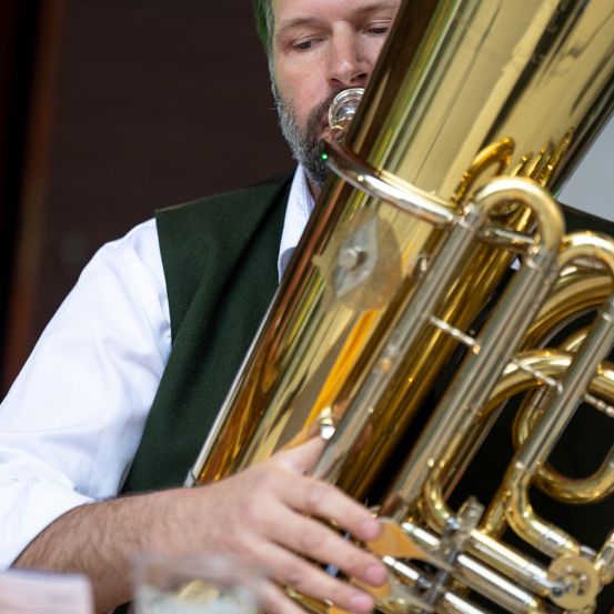 Ein Mann spielt eine große Messingtuba und konzentriert sich auf seine Darbietung, mit einem Glas Wasser in der Nähe.
