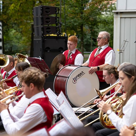 Ein Blasorchester spielt im Freien. Die Musiker sind in roten Westen und weißen Hemden gekleidet. Sie spielen verschiedene Instrumente, darunter Trompeten, Posaunen und Trommeln. Die Szene spielt in einem Park mit Bäumen und einem Gebäude im Hintergrund.