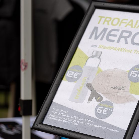 Ein Werbeplakat für Trofaiach-Merchandise bei einem StadtPARKfest-Event, mit einer Wasserflasche, einer Kappe und einem Ventilator, Preisen in Euro aufgeführt.