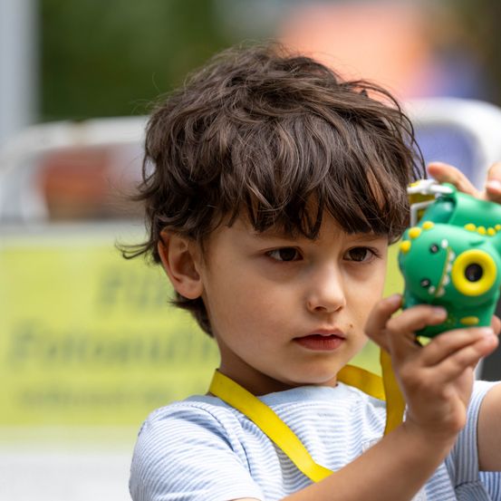 Ein junger Junge mit braunen Locken hält eine grüne Dinosaurier-Spielzeug in den Händen und sieht ernst aus. Er trägt ein gelbes Namensschild.