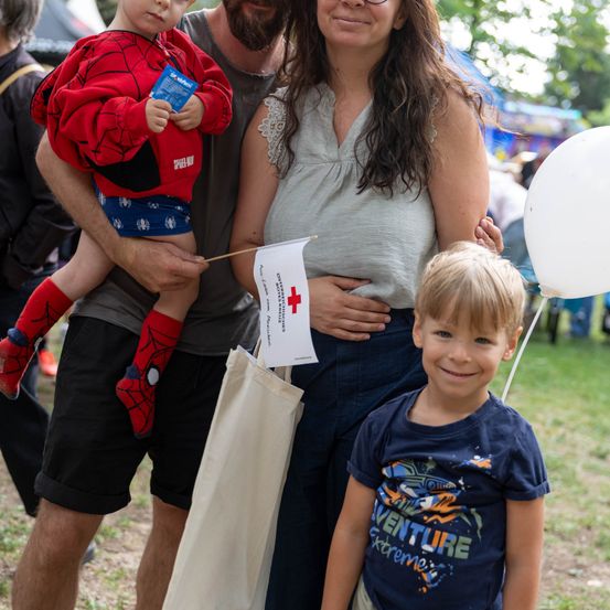Eine vierköpfige Familie steht bei einer Outdoor-Veranstaltung zusammen. Ein Junge in einem Spider-Man-Kostüm wird von einem Mann gehalten, während eine Frau eine Flagge mit einem roten Kreuz hält. Ein anderer Junge lächelt davor.