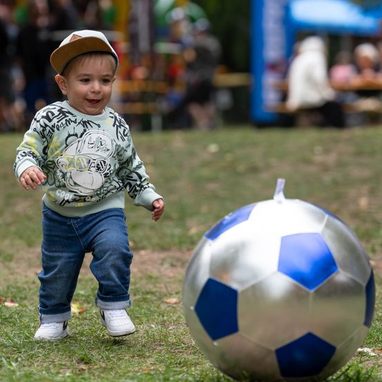 Ein kleiner Junge in einer Kappe und Jeans spielt mit einem großen, glänzenden Fußball auf einem Rasenfeld.