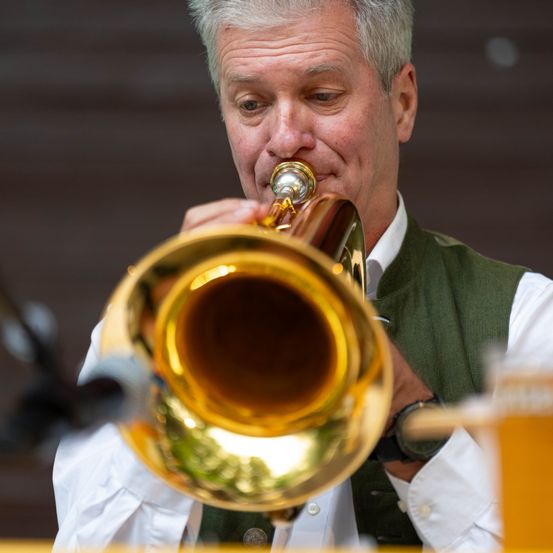 Ein Mann in einem weißen Hemd und einer grünen Weste spielt eine goldene Trompete und konzentriert sich auf seine Darbietung.