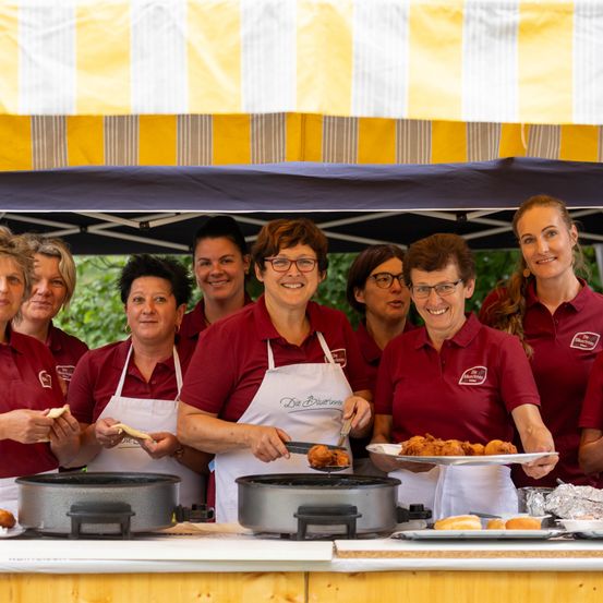 Eine Gruppe von Frauen in roten Hemden und weißen Schürzen lächelt und posiert für ein Foto vor einem Essensstand. Sie halten Essensartikel in den Händen und stehen unter einem gelb-weiß gestreiften Zelt.