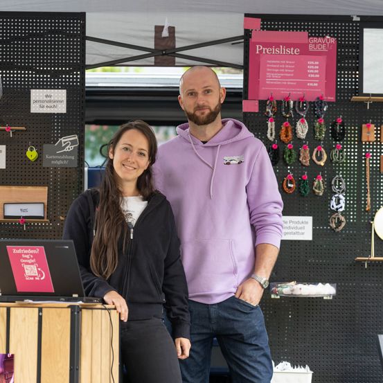 Ein Mann und eine Frau stehen lächelnd vor einem Stand mit einem Laptop. Der Mann trägt eine lila Kapuzenjacke und die Frau eine schwarze Jacke. Hinter ihnen ist eine Preisliste an einer Wand mit verschiedenen Artikeln angeheftet.
