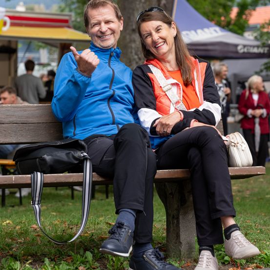 Ein Mann und eine Frau sitzen auf einer Bank, lächeln und posieren für ein Foto. Der Mann trägt eine blaue Jacke und die Frau einen orangefarbenen Weste. Sie haben Taschen auf der Bank und sind von einem Park mit Zelten und Menschen umgeben.