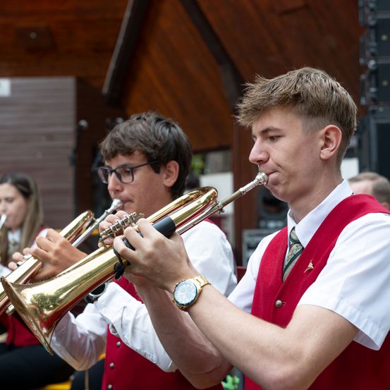 Zwei junge Männer in roten Westen und weißen Hemden spielen Trompeten in einer Außenumgebung, wahrscheinlich bei einem Konzert.