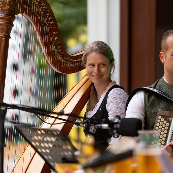 Eine Frau spielt Harfe und ein Mann Akkordeon. Sie stehen nebeneinander und lächeln. Sie scheinen auf der Bühne zu spielen.