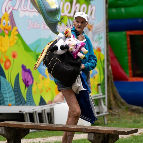 Eine ältere Frau mit einem Rucksack voller Plüschtiere geht an einem bunten Hintergrund mit Luftballons und einem Hüpfhaus vorbei.