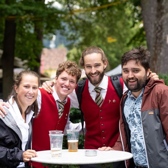 Vier Personen, zwei Männer und zwei Frauen, lächeln und posieren für ein Foto in einem Park. Sie tragen rote Westen und halten Gläser mit Bier.