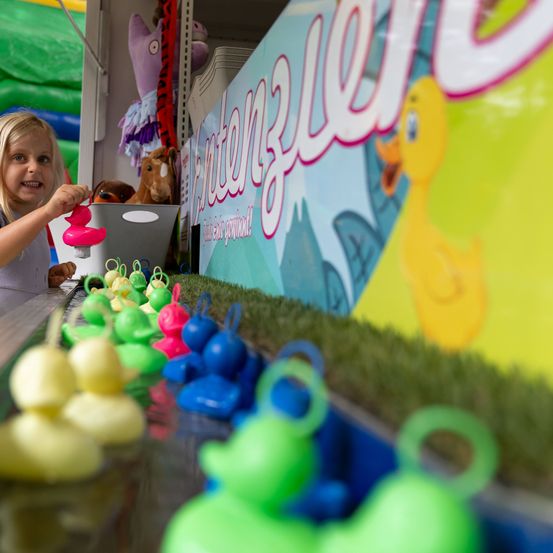 Ein kleines Mädchen spielt mit einem ententhematischen Spiel auf einem Jahrmarkt, umgeben von bunten Enten.