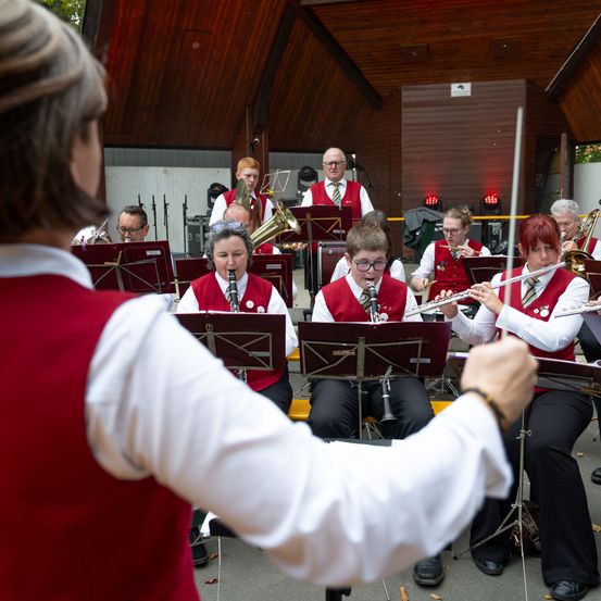 Ein Orchester spielt im Freien mit einem Dirigenten. Die Musiker spielen verschiedene Instrumente, darunter Trompeten, Posaunen und Flöten. Der Ort ist ein hölzerner Pavillon.