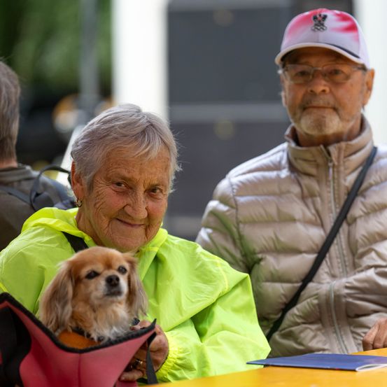 Eine ältere Frau mit grauem Haar hält einen kleinen Hund in einem roten Tragekorb. Sie trägt einen leuchtend gelben Regenmantel. Neben ihr steht ein älterer Mann mit Brille und Baseballkappe.