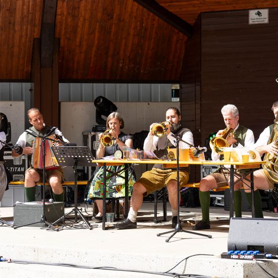 Eine Gruppe von Musikern tritt auf der Bühne auf, einer spielt Akkordeon, andere Trompeten und eine Frau spielt Flöte. Sie sitzen an einem Tisch mit Getränken und einem Mikrofon. Die Umgebung ist im Freien mit Holzwänden.
